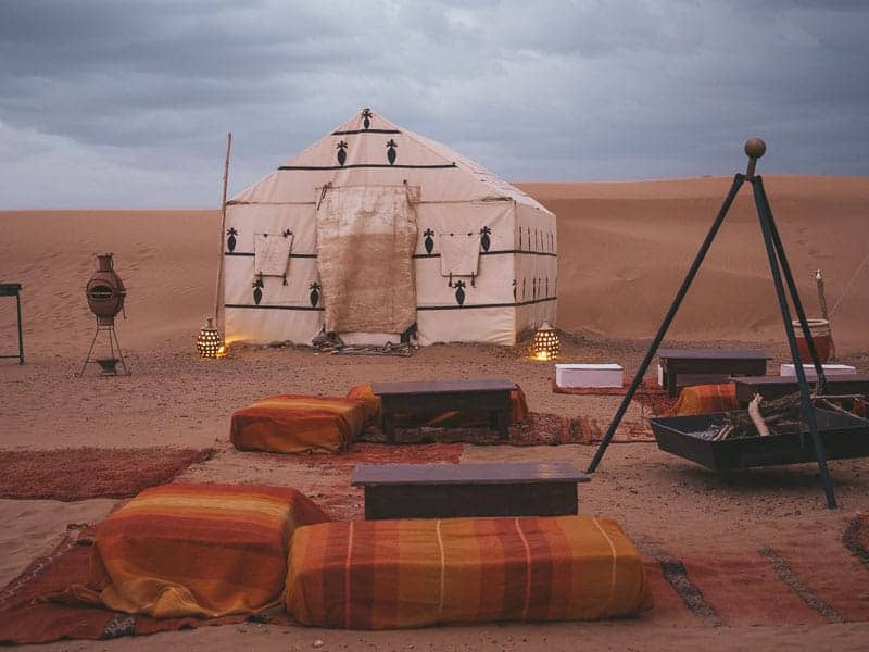 Sahara Desert glamping experience with white canvas tent and fire pit surrounded by Moroccan rugs and cushions.